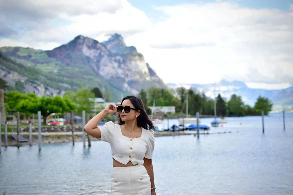 Young woman in a stylish white outfit poses by a serene lake with mountains in the background on a sunny day.