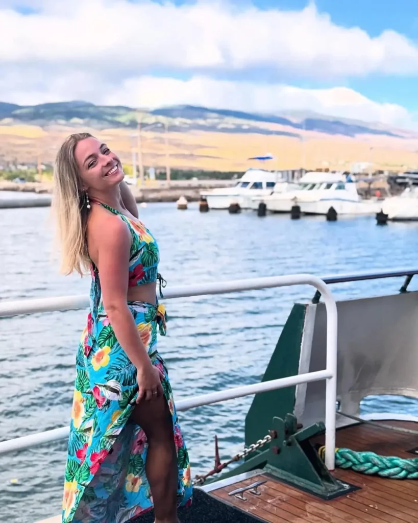 Smiling woman in a colorful floral dress poses by the water, with boats and mountains in the background on a sunny day.