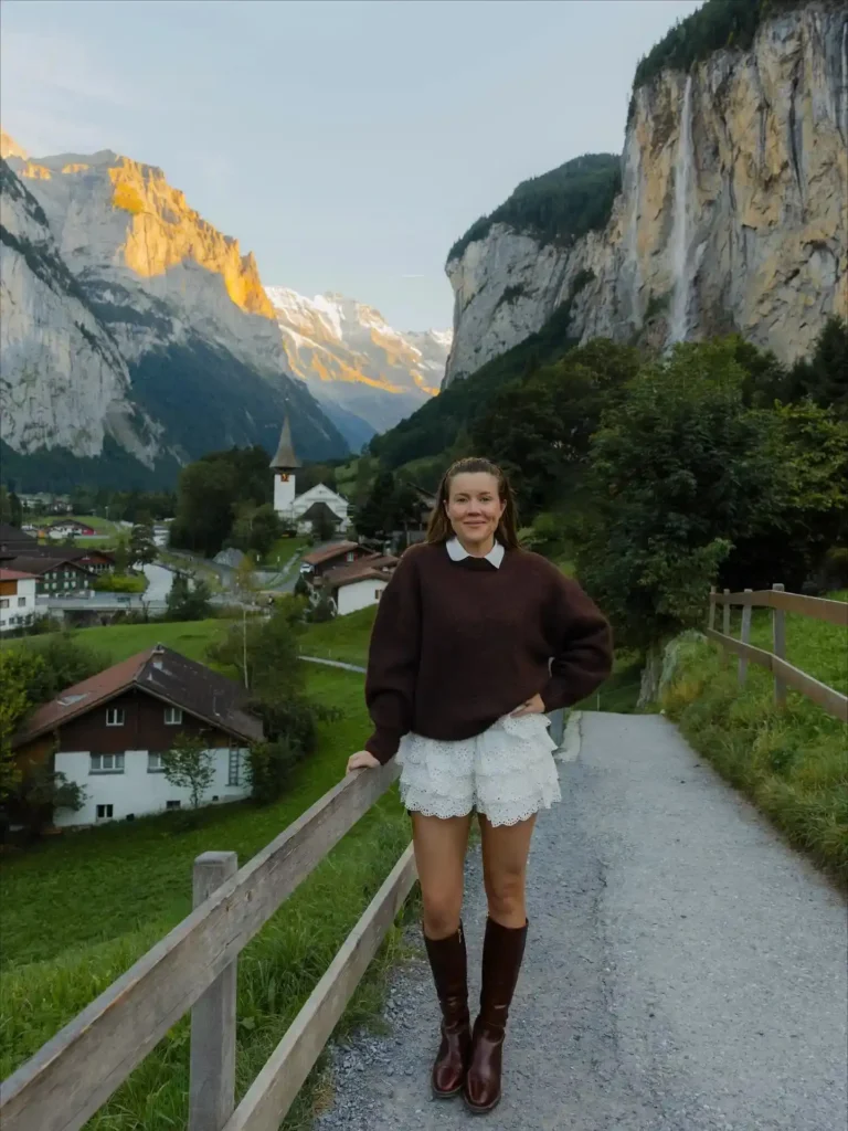 A woman in a brown sweater and white shorts poses by a scenic mountain village at dusk.
