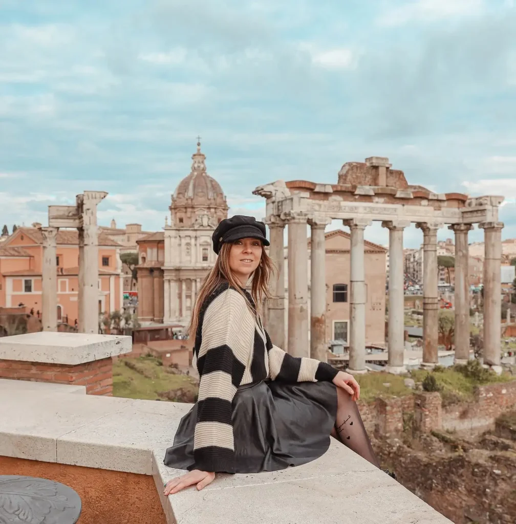 Young woman in stylish outfit sitting near ancient Roman ruins in a scenic outdoor setting with cloudy sky.