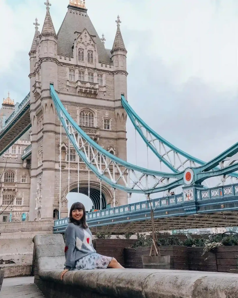 Young woman sitting by the Thames with Tower Bridge in the background, showcasing London's iconic architecture.