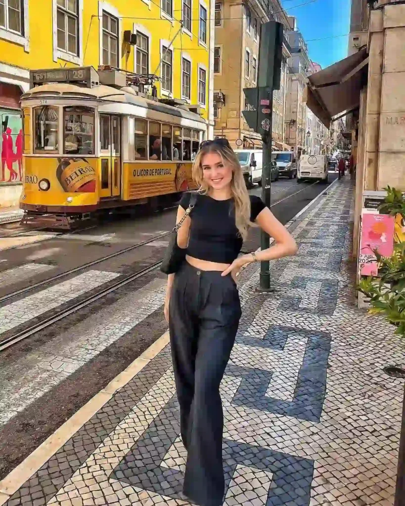 Woman in black crop top and pants posing on a Lisbon street with a vintage yellow tram and vibrant architecture.