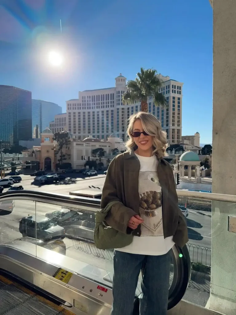 Young woman in sunglasses smiles near an escalator with Las Vegas skyline in the background on a sunny day.