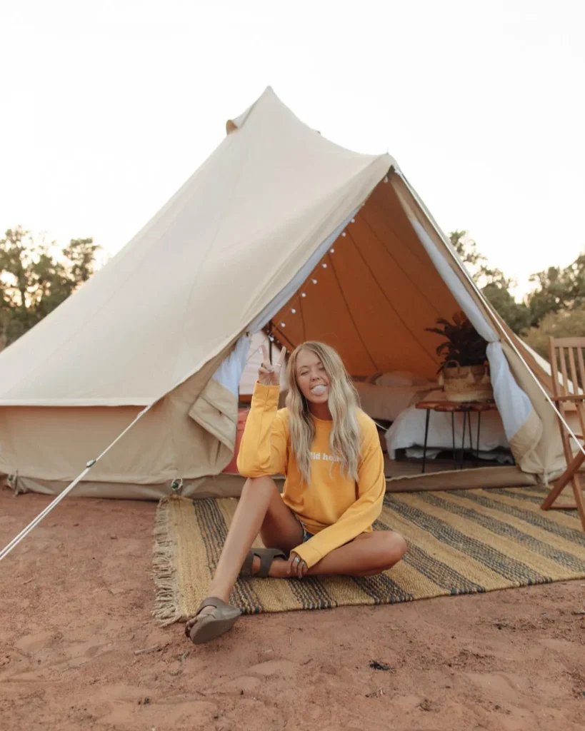 Young woman with long blonde hair sitting outside a glamping tent, giving a peace sign and enjoying nature.