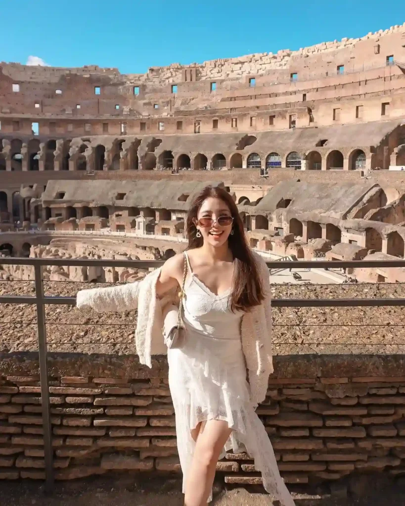 Young woman in a white dress poses near the Colosseum in Rome, showcasing the historic architecture and blue sky.