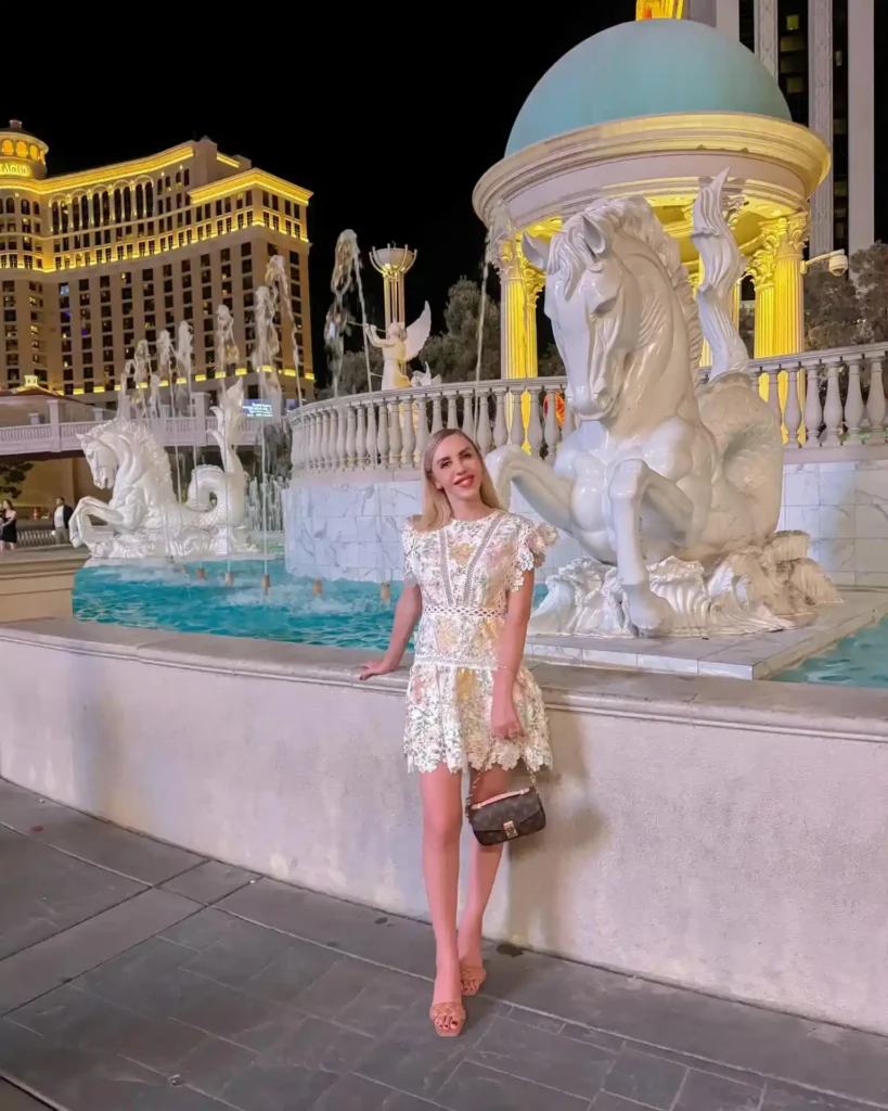 Young woman in a lace dress poses by a white horse statue at night in Las Vegas, with illuminated buildings in the background.