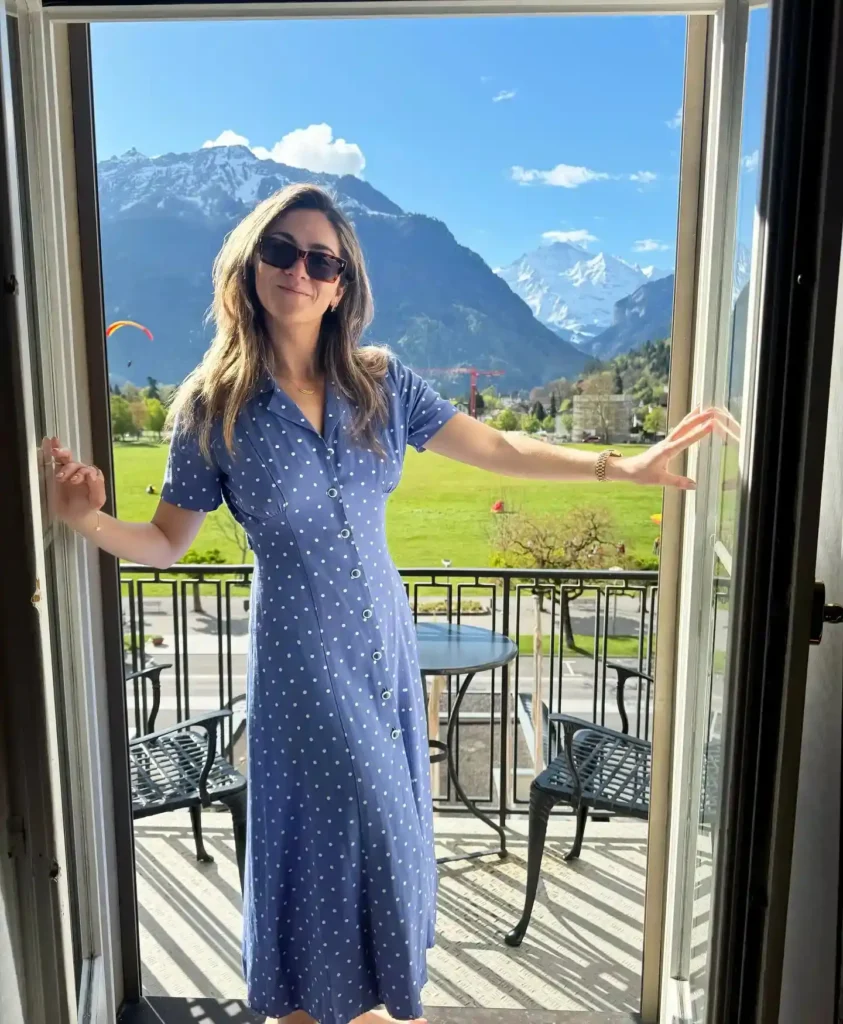 Woman in a polka dot dress stands at a balcony, showcasing a stunning mountain view in a sunny landscape.