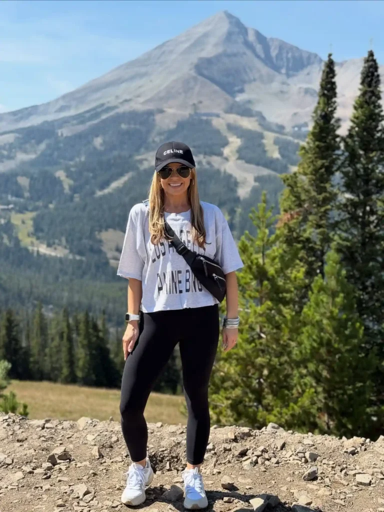 Smiling woman in a graphic t-shirt and leggings stands on a mountain trail with pine trees and peaks in the background.