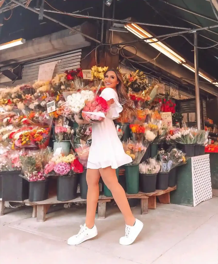 Woman in a white dress joyfully holding flowers at a vibrant flower market filled with colorful blooms.