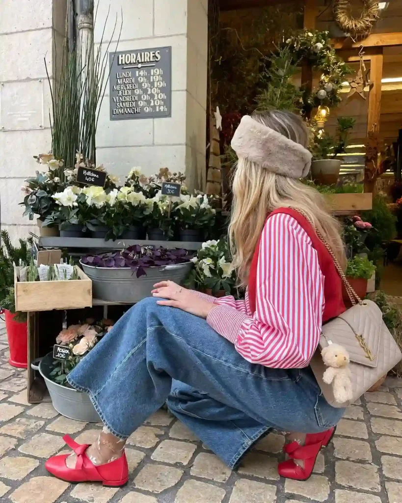 A woman in a red and white striped shirt kneels by a flower shop, surrounded by potted plants and a sign displaying hours.