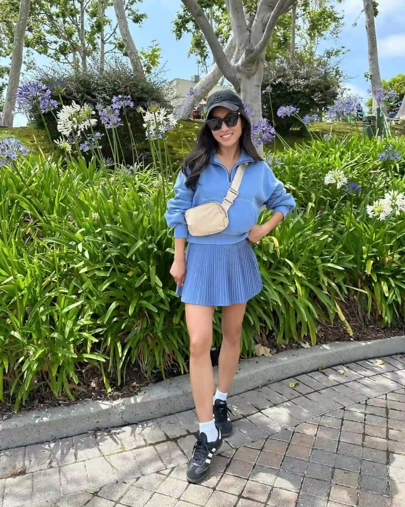 Woman smiling in a blue outfit with a pleated skirt, standing among lush greenery and flowers on a sunny day.