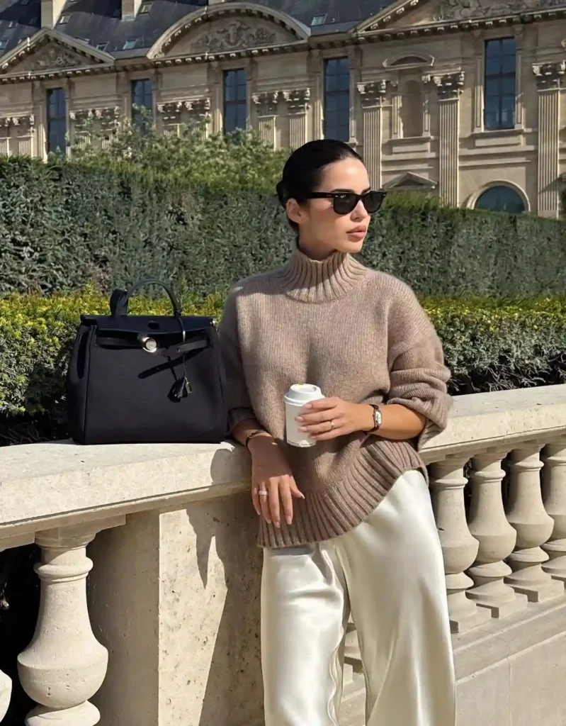 Fashionable woman in a beige sweater and silk pants, holding a coffee cup, posing in front of elegant architecture.
