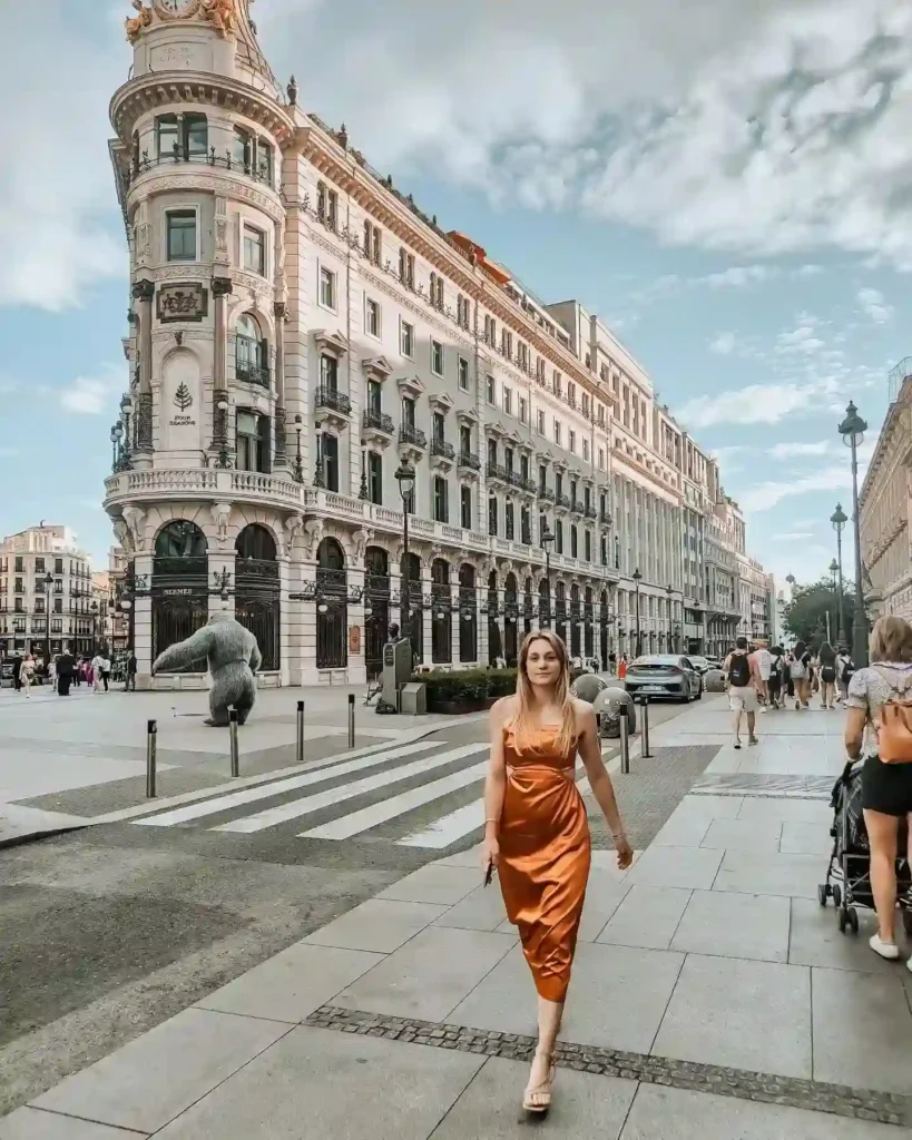 A woman in an orange dress walks down a busy street with beautiful architecture and a cloudy sky in the background.