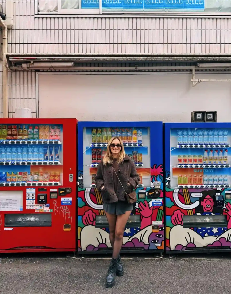 A woman in a stylish outfit stands between colorful vending machines in an urban setting, smiling and enjoying the day.
