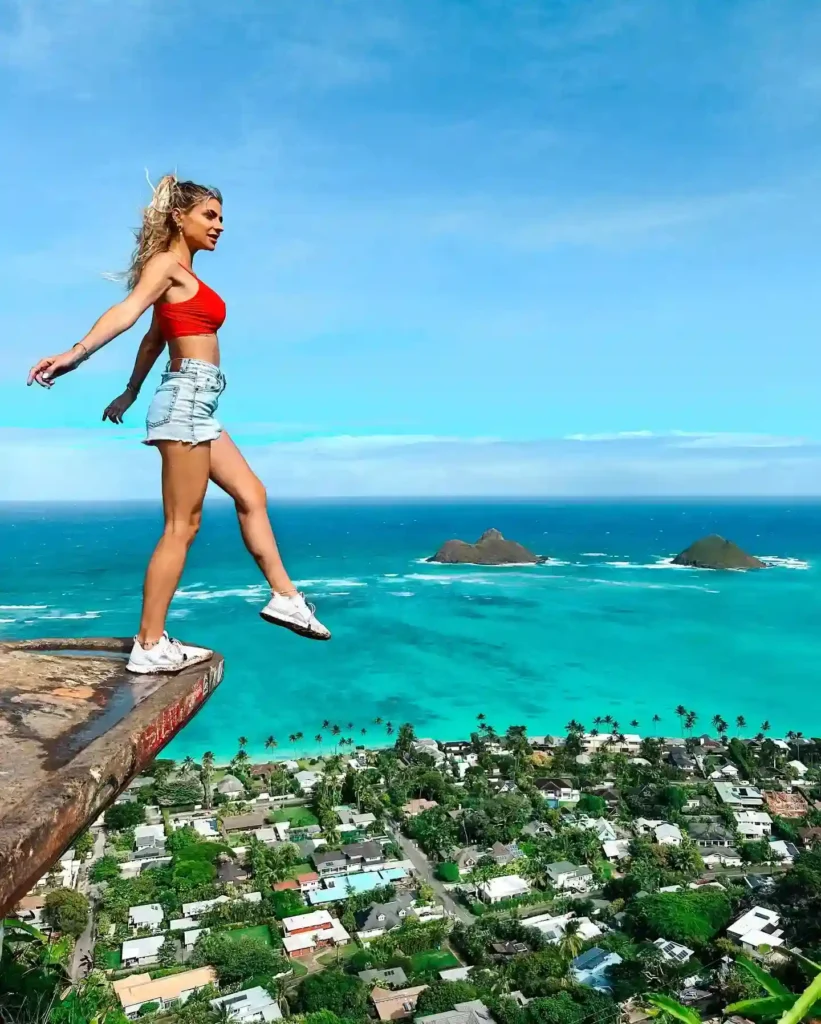 A woman in a red crop top walks confidently along a cliff edge overlooking turquoise ocean and scenic landscape.