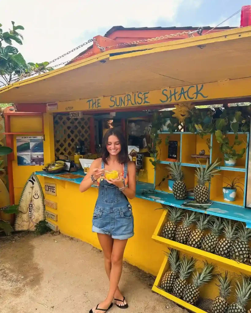 Smiling woman holding fresh fruit outside The Sunrise Shack, a vibrant yellow stand with pineapples and tropical decor.