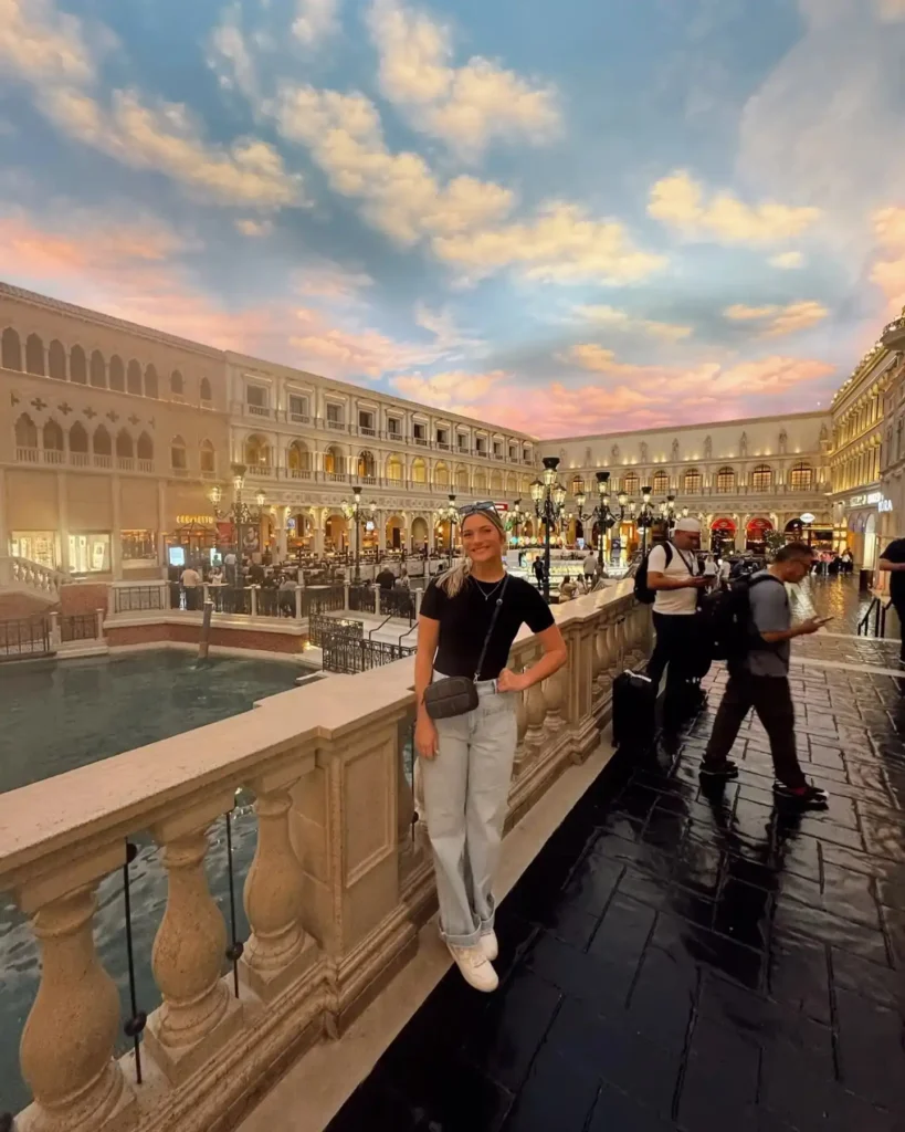 A woman poses by a canal in The Venetian, Las Vegas, as vibrant clouds fill the sunset sky behind her.