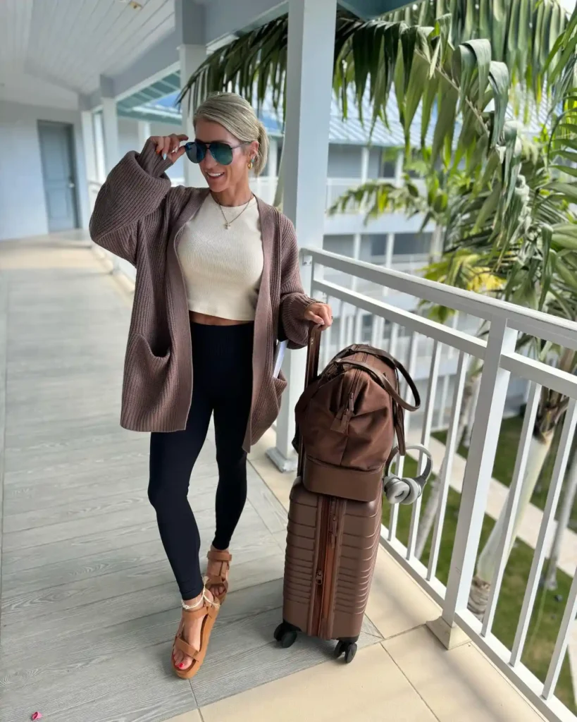 Woman in stylish outfit poses with suitcase and bag on a balcony surrounded by palm trees, ready for travel.