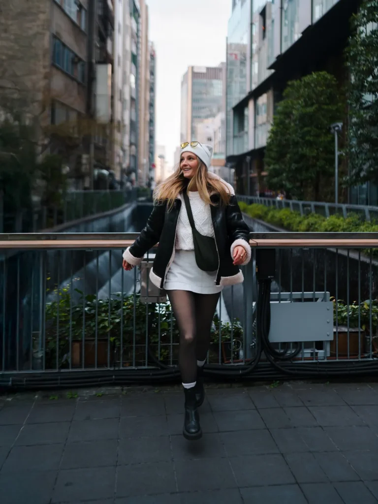Young woman in stylish winter outfit joyfully walking in an urban area, surrounded by modern buildings and greenery.