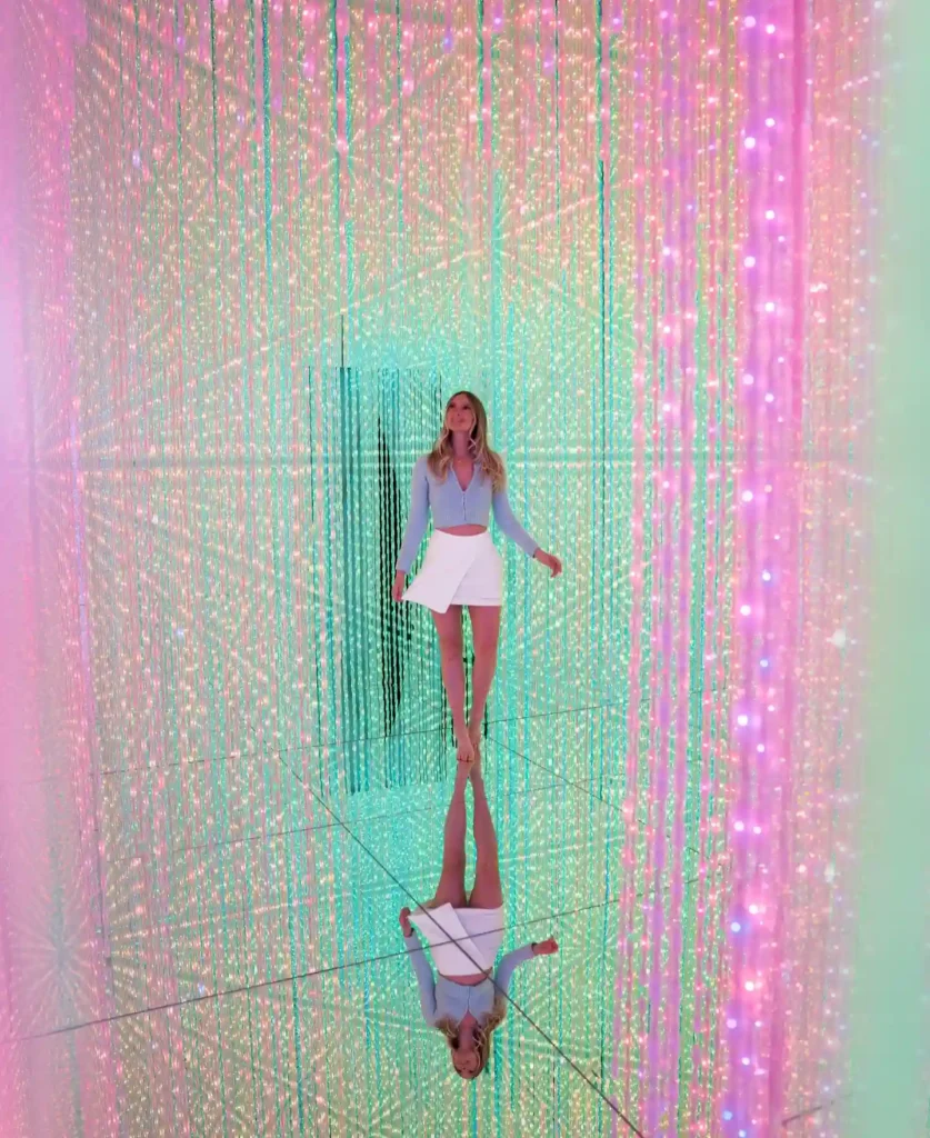 A woman in a stylish outfit stands in a colorful, mirrored space filled with sparkling LED lights.
