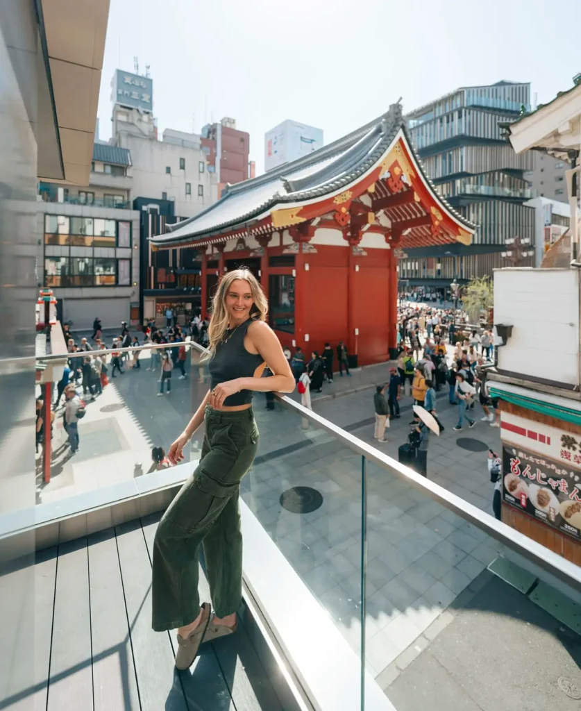 A young woman poses on a balcony with a traditional Japanese temple and bustling street scene in the background.