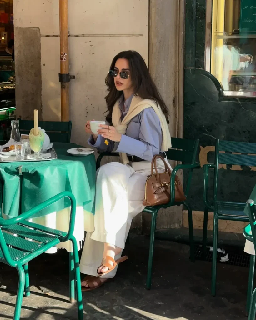 Young woman sitting at a café table, enjoying a coffee in stylish attire with a sweater draped over her shoulders.