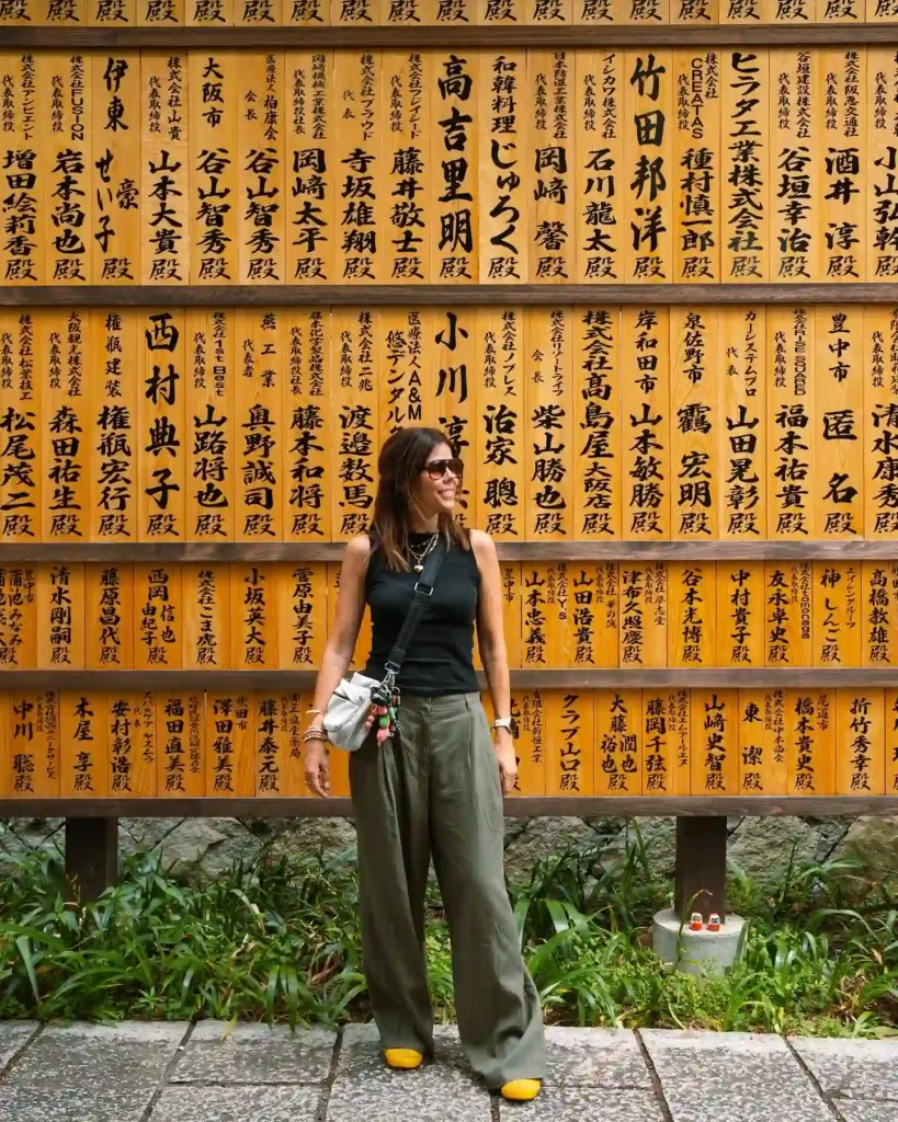 Woman standing in front of a traditional Japanese wooden signboard filled with names, wearing a black top and green pants.