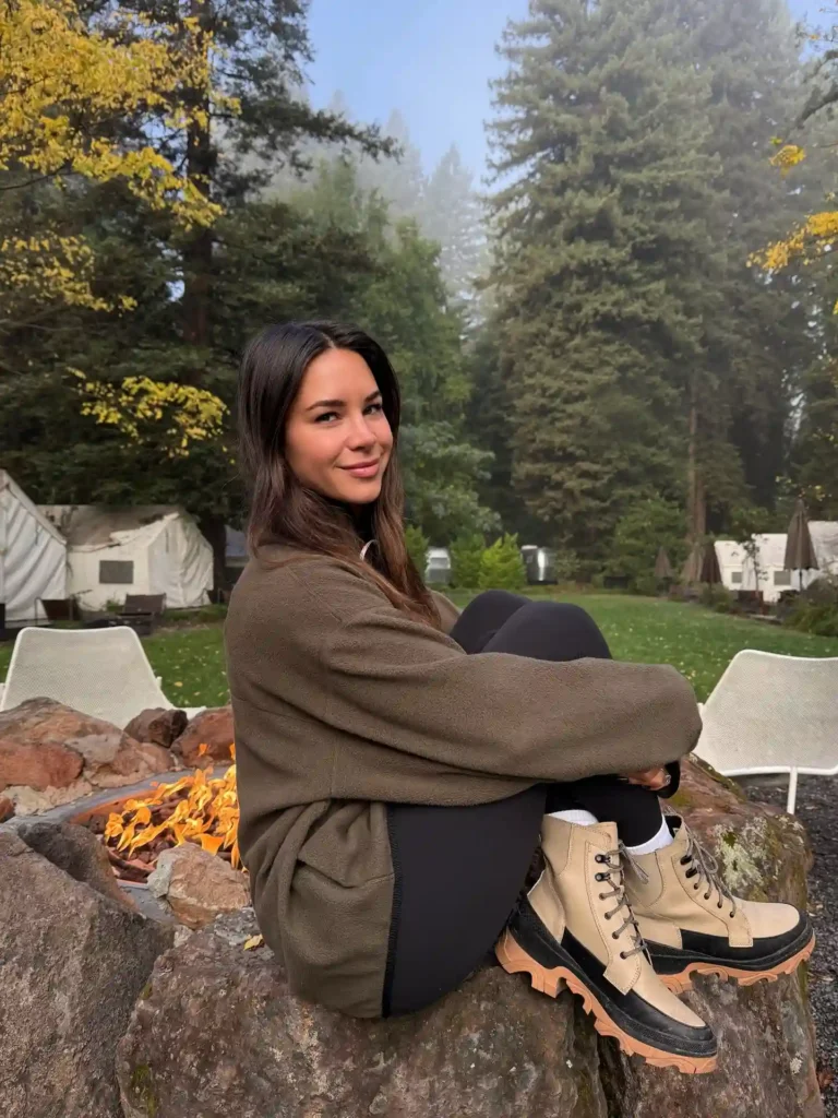 Young woman sitting on rocks by a campfire in forest, wearing casual hiking attire and smiling at the camera.