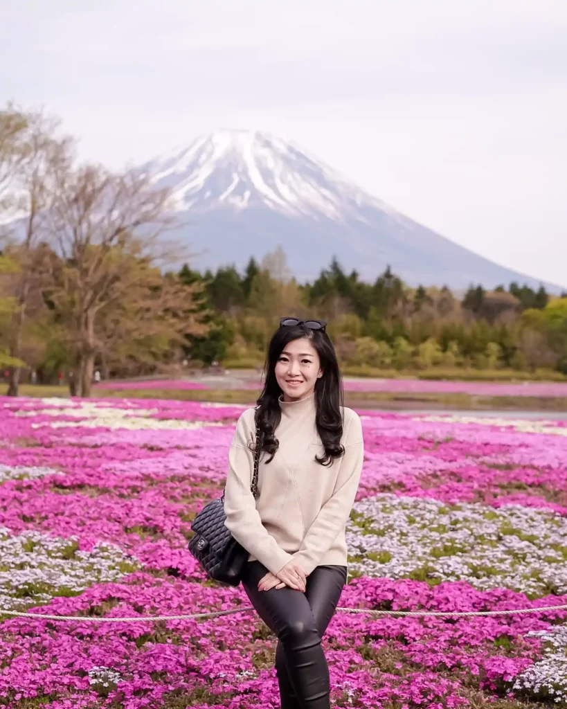 Woman in a light sweater poses amid vibrant pink flowers with Mount Fuji in the background on a clear day.