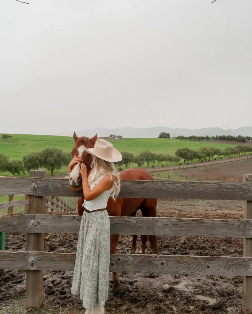 Woman in a hat affectionately interacting with a horse by a wooden fence in a lush countryside setting.