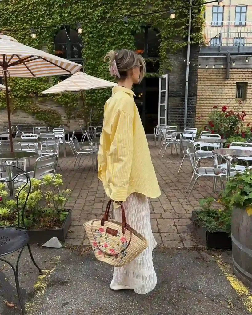 A woman in a yellow shirt and white pants holds a floral bag, standing in a charming outdoor café setting.
