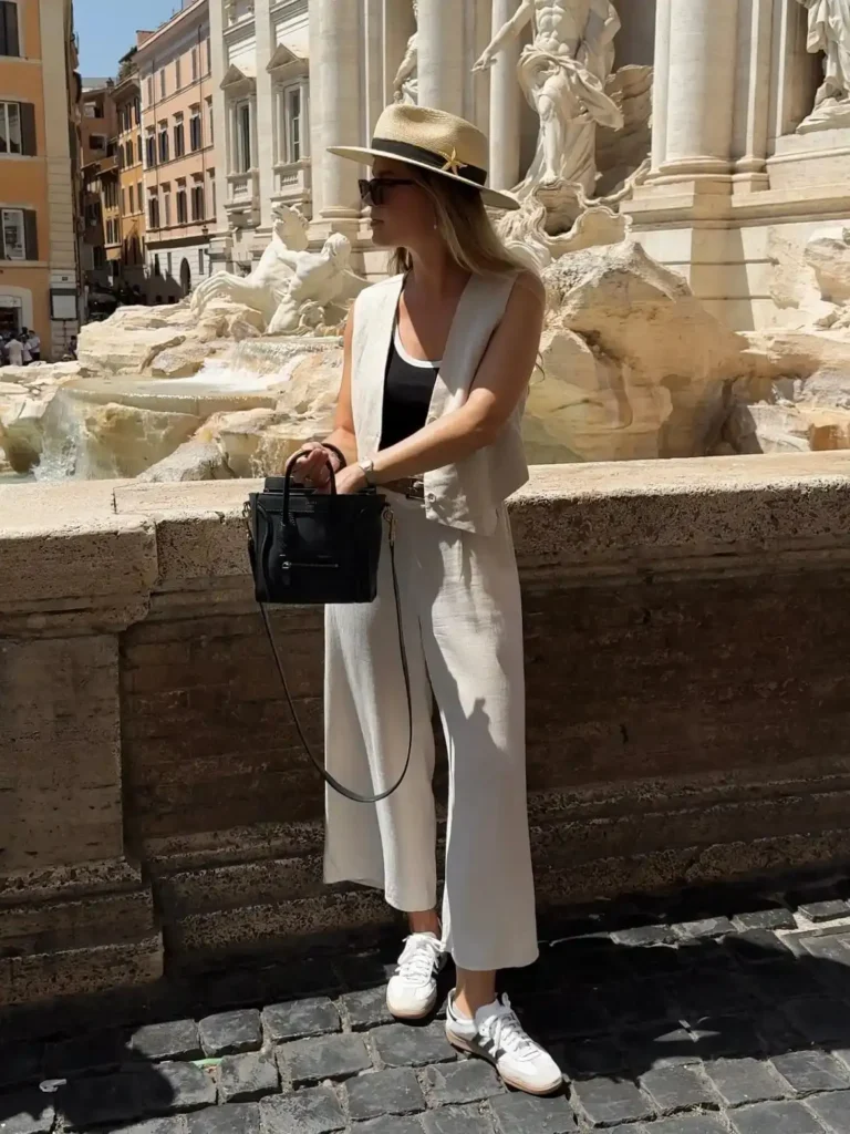 Stylish woman in a light outfit and hat stands by a fountain in Rome, enjoying a sunny day in the historic city.