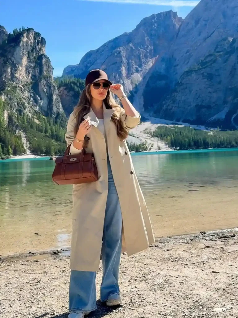 A woman in a beige coat and sunglasses stands by a serene lake, surrounded by majestic mountains under a clear blue sky.