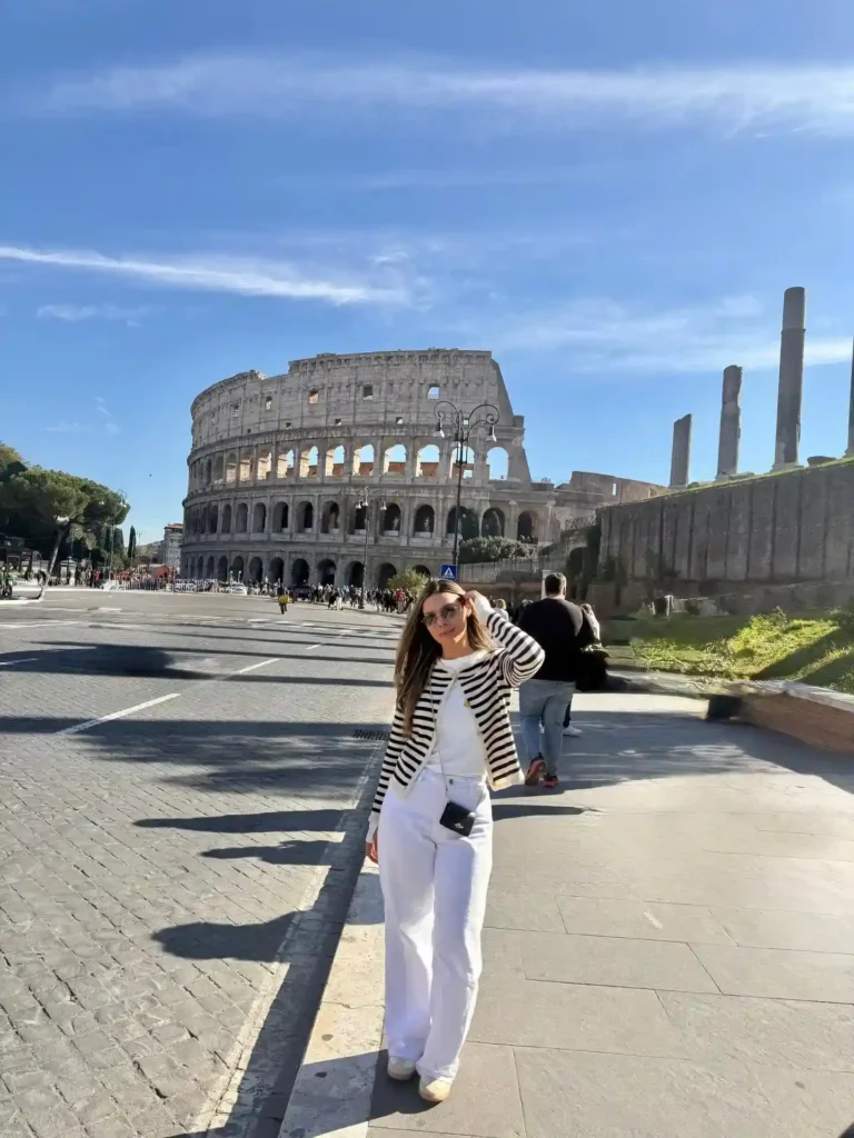 Woman in stylish outfit poses near the Colosseum in Rome on a sunny day, with tourists in the background.