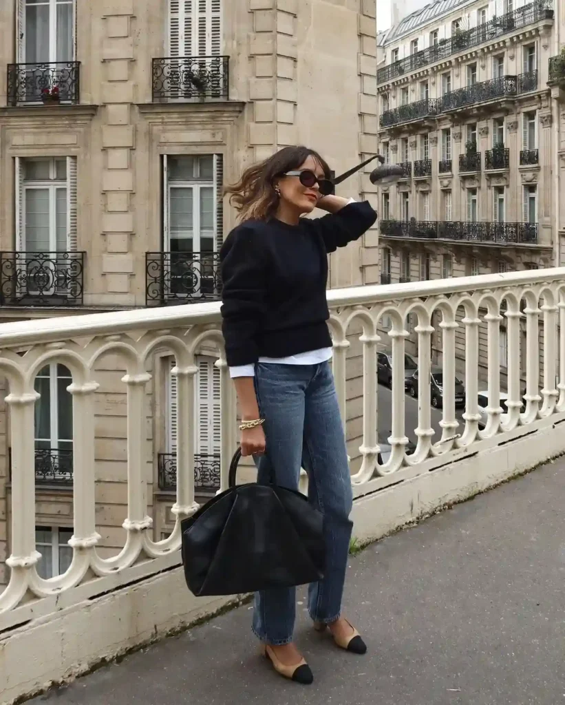 Chic woman in sunglasses wearing a black sweater and jeans, posing on a Parisian balcony with stylish black handbag.