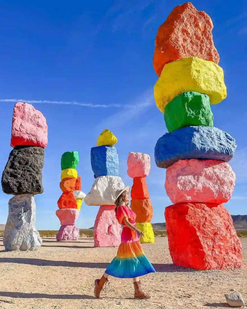 A woman in a colorful dress walks past vibrant rock formations under a clear blue sky at the Seven Magic Mountains.