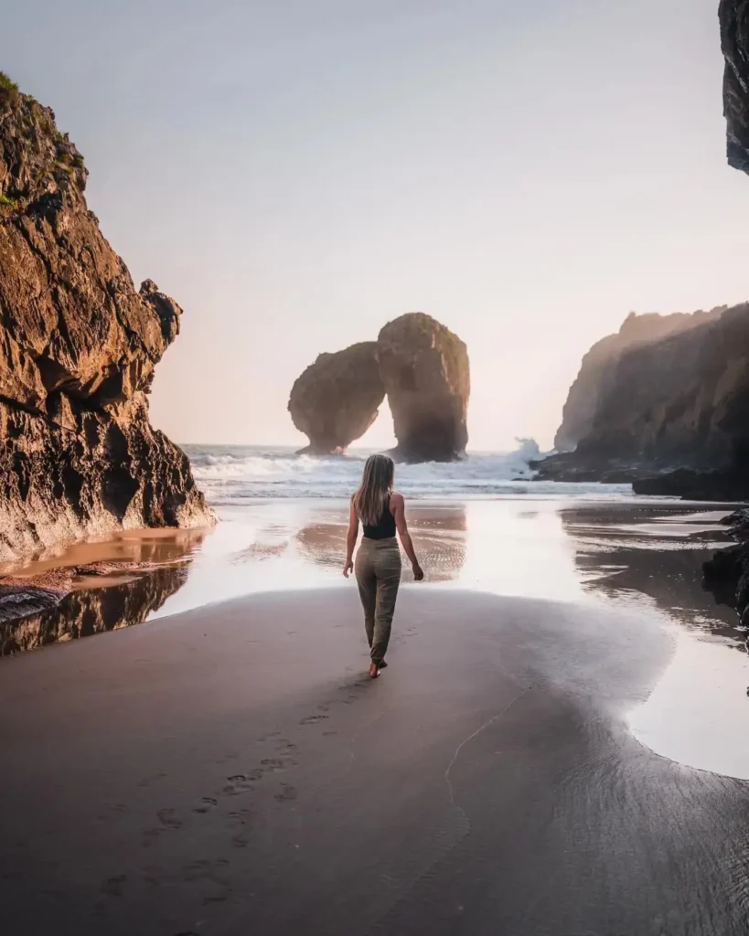 Woman walking on a sandy beach at sunset, flanked by rocky cliffs and natural arches in the background, reflecting tranquility.