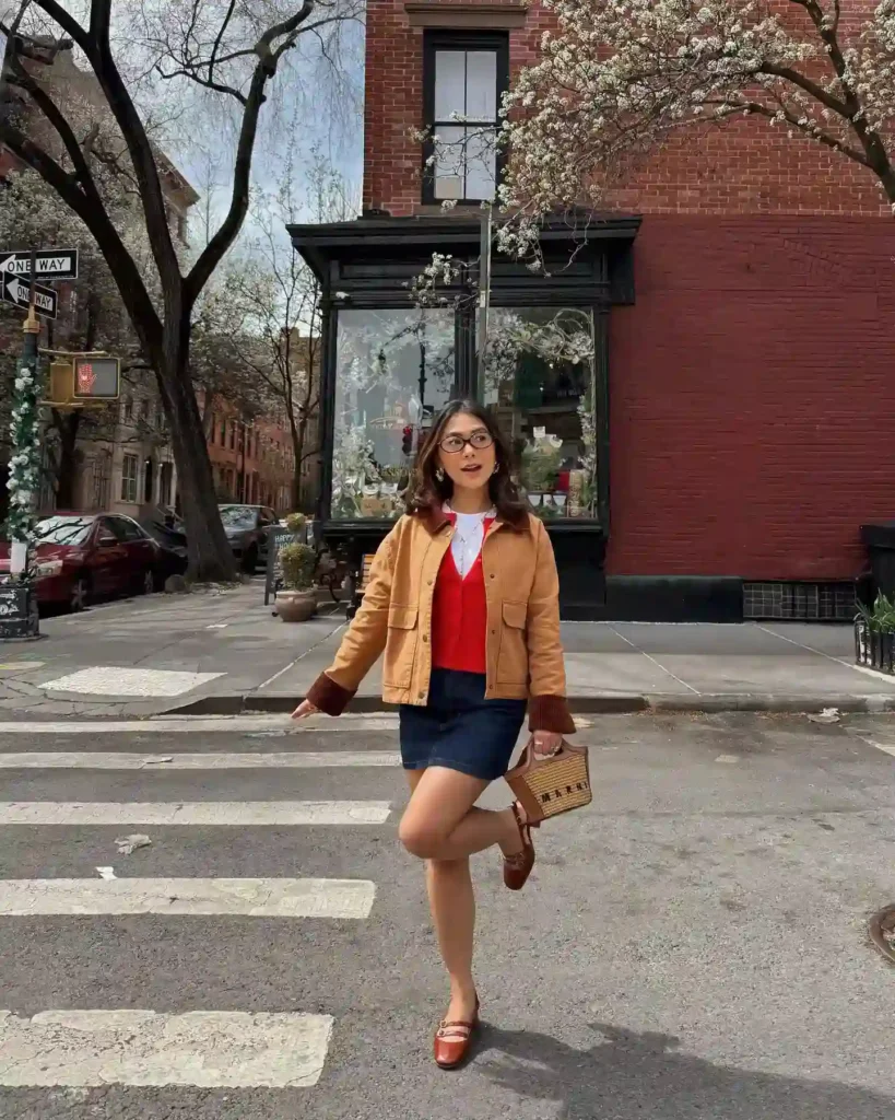Young woman in stylish outfit crossing a city street, surrounded by blooming trees and vintage architecture.