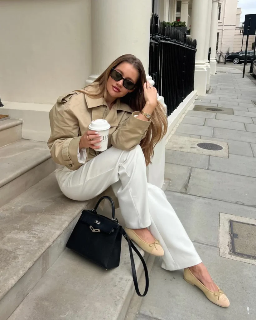 Stylish woman in a beige jacket and white pants, sipping coffee while sitting on steps with a black handbag.