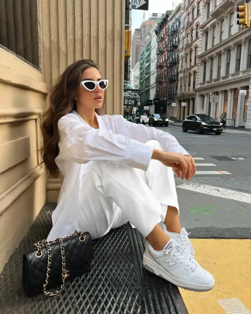 Fashionable woman in white outfit and sunglasses sitting on a city sidewalk, showcasing style and urban scenery.