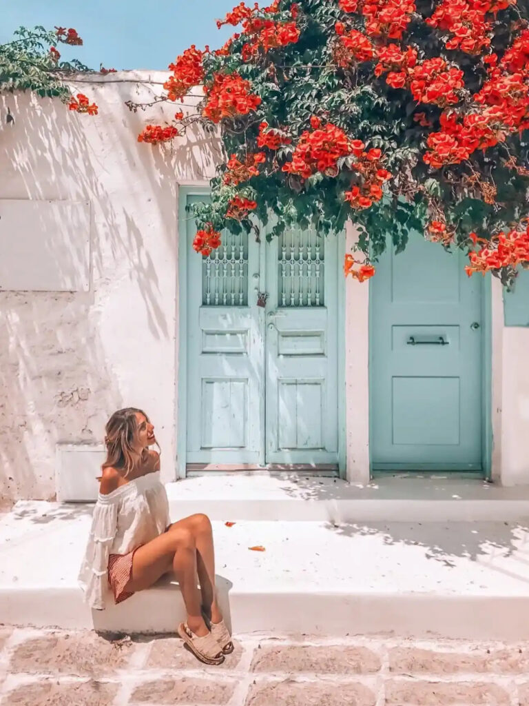 Woman in a white off-shoulder top relaxes on steps by blue doors, surrounded by vibrant red flowers in a sunny location.