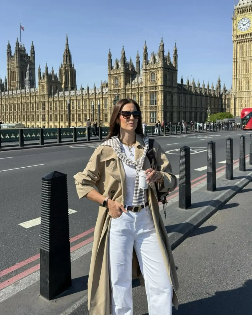 Woman in a beige trench coat stands in front of the Houses of Parliament in London, enjoying a drink on a sunny day.