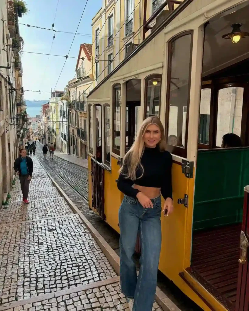 Woman in casual attire standing beside a vintage yellow tram on a cobblestone street in a scenic city.