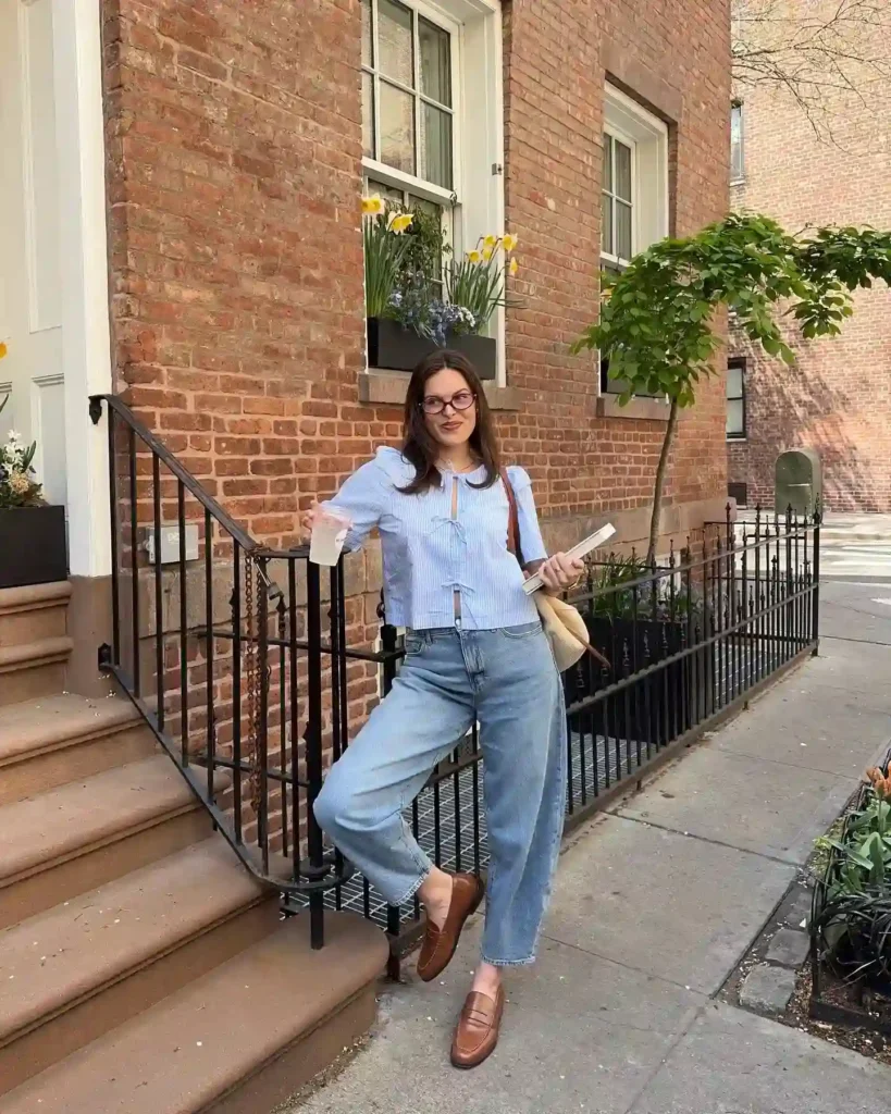 Young woman in casual outfit poses on steps of a brownstone, holding a drink and book, surrounded by flowers.