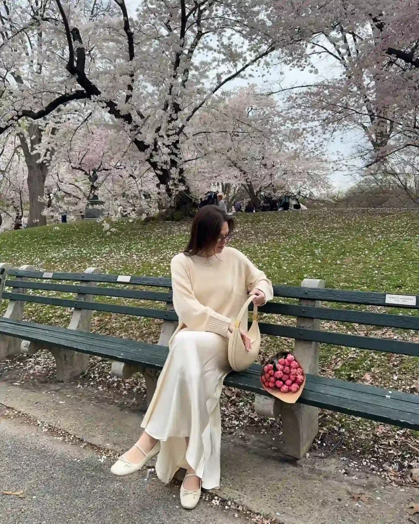 Woman in a cream sweater and white skirt sits on a park bench, surrounded by blooming cherry blossom trees and flowers.