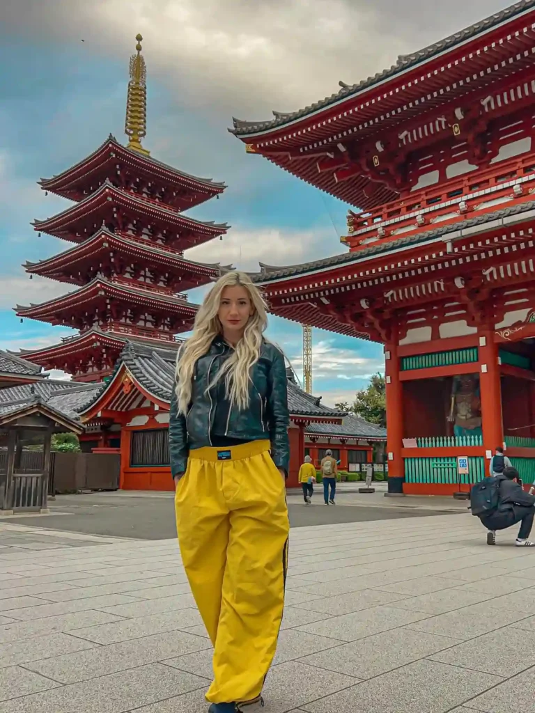 Woman in stylish yellow pants and leather jacket poses in front of traditional Japanese pagoda and temple architecture.