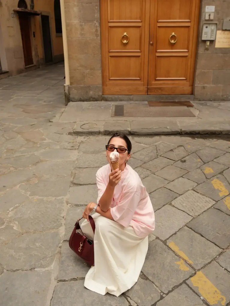 Young woman enjoying ice cream while sitting on cobblestone street, dressed in a pink shirt and white skirt.