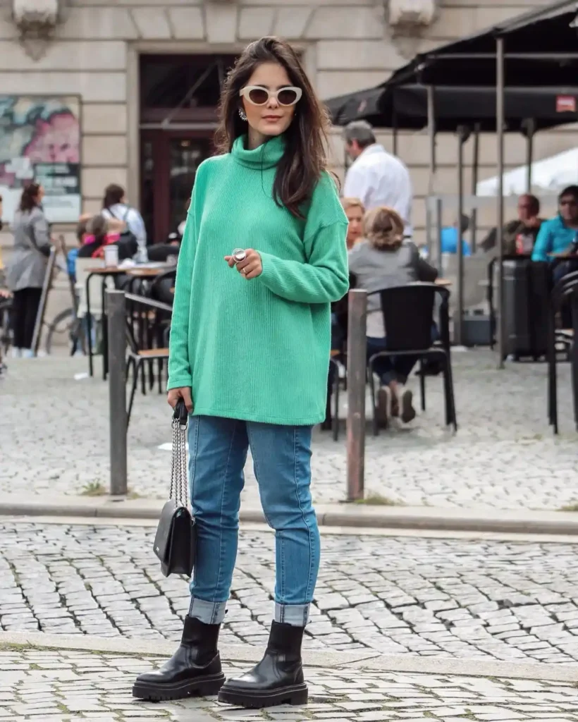 Young woman in a mint green sweater and black boots poses stylishly on a cobblestone street. Casual urban fashion look.