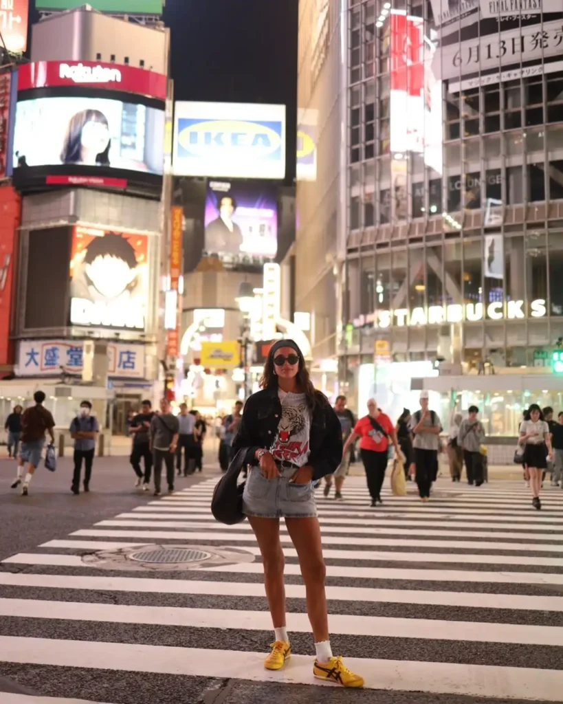 A woman poses in stylish attire at a busy Shibuya crossing with vibrant city lights and crowds in the background.