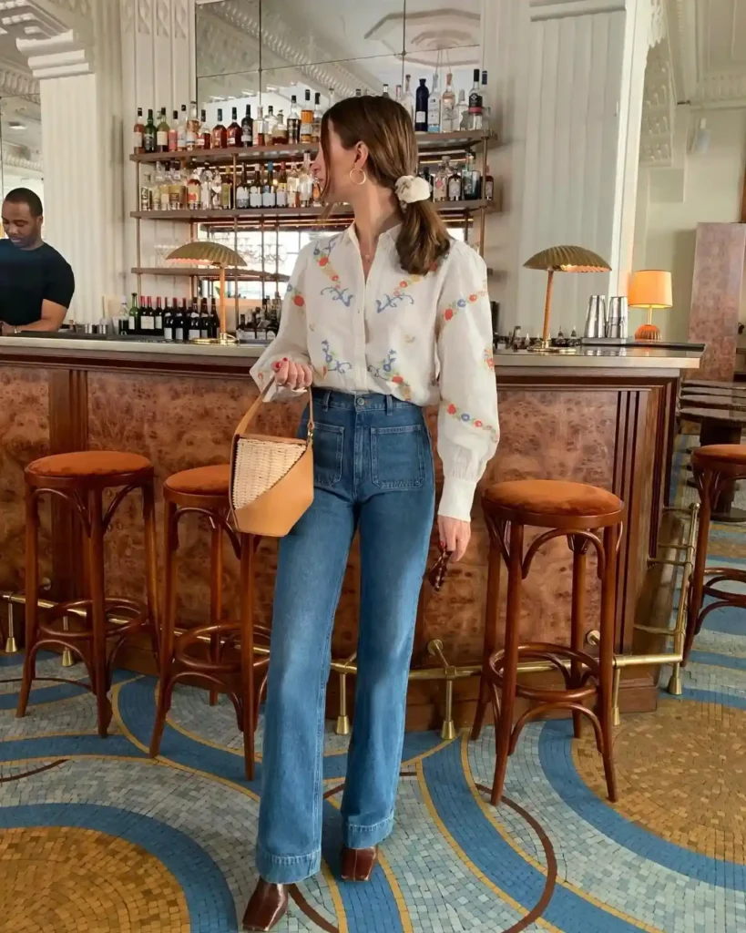 Woman in stylish outfit featuring a floral blouse and high-waisted jeans, standing at a chic bar setting.